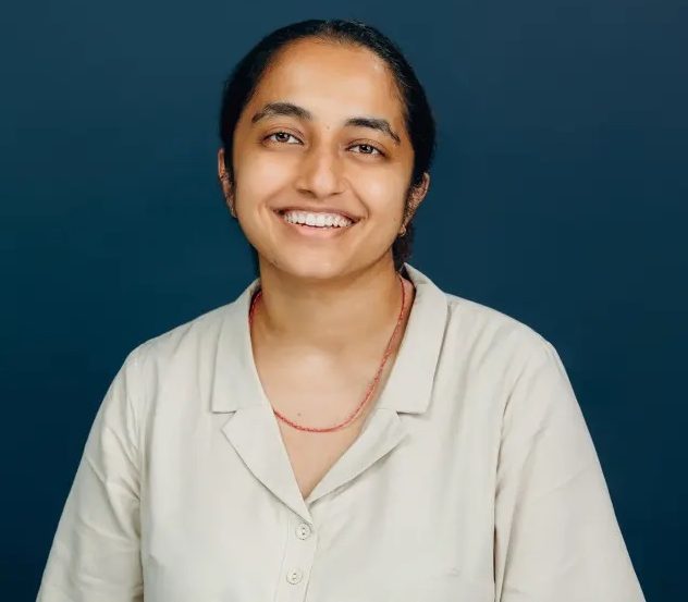 Kim, a brown person with long black hair, wears a cream collared shirt. They are smiling at the camera, and their hair is pulled back.