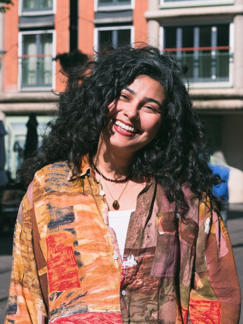 A woman with dark curly hair and a patterned brown shirt stands in a sunny street, smiling.