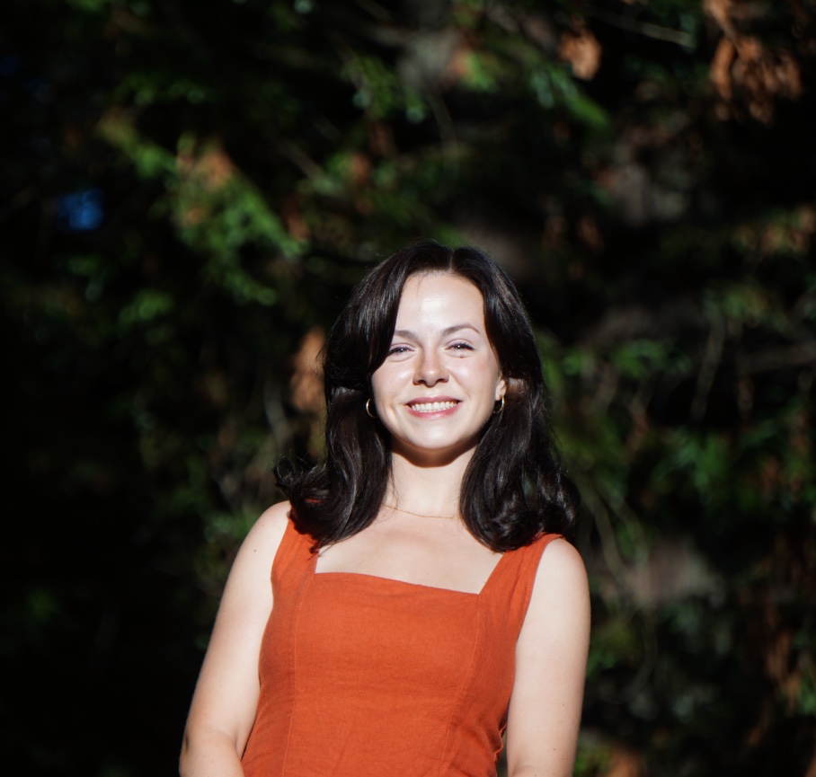 A woman with shoulder-length dark hair smiles at the camera while standing outdoors in bright sunlight. They are wearing a sleeveless burnt orange dress, small hoop earrings, and a delicate necklace. The background is softly blurred, with dark green foliage from redwood trees.