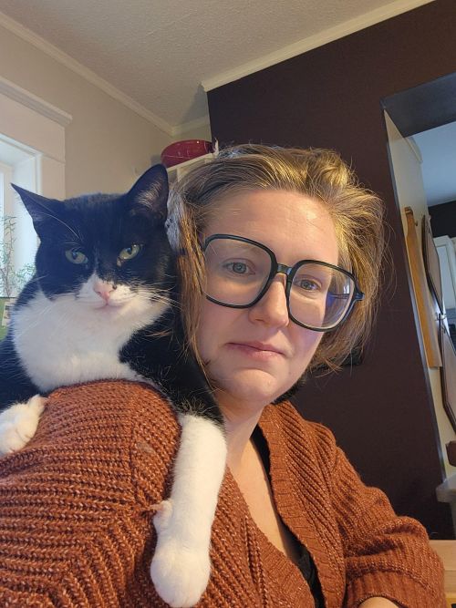 A white female with short reddish brown hair and large blue-framed glasses. She is wearing an orange cardigan and looking at the camera. There is a black and white cat draped over her shoulder with a very pink nose, also staring at the camera.